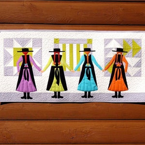 Könnte beinhalten: Ein gequiltetes Wandbehang mit vier Figuren in schwarzen Kleidern mit bunten Röcken. Die Figuren halten sich an den Händen und haben rote Haare und schwarze Hüte. Der Hintergrund ist ein weißer Stoff mit einem geometrischen Muster.