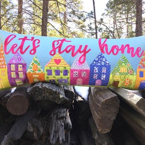 以下が含まれることがあります： 青い背景にピンクの文字で「Let's stay home」と書かれた長方形の枕。枕には、さまざまな模様のカラフルな家が並んでいます。