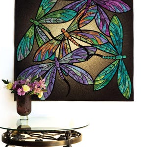 Op de afbeelding: Een wanddecoratie met een kleurrijk quilt met een ontwerp van vijf libellen. De libellen zijn gemaakt van verschillende gekleurde stofstukken en hebben een glas-in-loodlook. De achtergrond is donkerbruin.