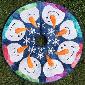 May include: A round, blue Christmas tree skirt featuring smiling snowmen with orange carrot noses and colorful scarves. White snowflakes are scattered around the snowmen. The skirt has a center hole for the tree trunk.
