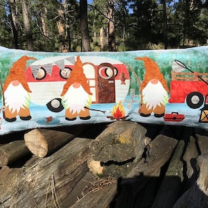 Op de afbeelding: Een lang decoratief kussen met een blauwe en groene achtergrond waarop een rode en witte camper, vier gnomes, een kampvuur en een rode wagen zijn afgebeeld. De gnomes dragen rode mutsen en hebben witte baarden. Het kussen ligt op een stapel houtblokken.