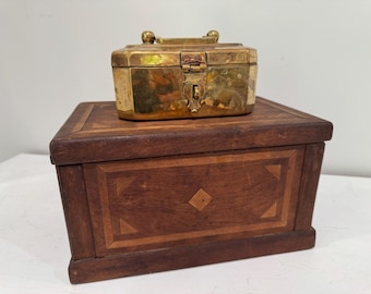May include: A small, antique brass box with a handle and clasp sits atop a larger wooden box. The wooden box features inlaid geometric patterns in a darker wood, creating a decorative contrast. The brass box has a tarnished appearance.