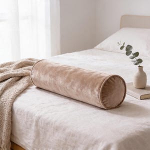 May include: A cylindrical bolster pillow made of soft, light beige velvet fabric with piped edges. The pillow is positioned horizontally on a bed covered with a white linen sheet and a textured, cream-colored knit throw blanket.