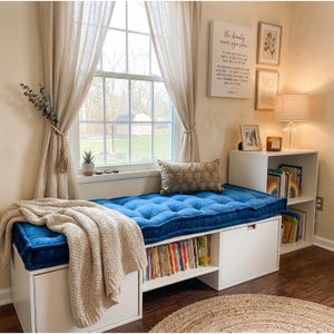 May include: A cozy window seat with a tufted blue cushion and a beige throw blanket. The seat is built into a white storage unit with open shelves filled with books. A decorative pillow and framed art complete the inviting reading nook.