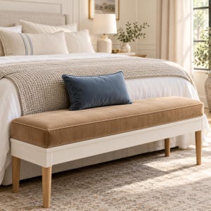 May include: A rectangular bench featuring a tan upholstered seat with piped edges, supported by a white wooden frame and four light-toned wooden legs. The bench is positioned at the foot of a bed, topped with a single rectangular blue pillow.