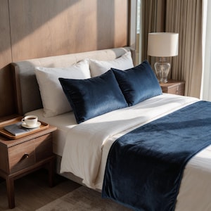 May include: A bed with white linens, two navy blue velvet pillows, and a matching bed runner. A wooden nightstand with a lamp and a tray with a cup and saucer are visible. The room has a neutral color palette.