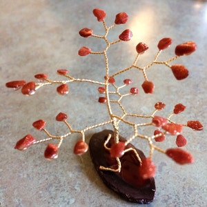 May include: A handcrafted wire tree sculpture with reddish-orange gemstone leaves. The tree's branches are made of gold-colored wire, and it is mounted on a dark brown, oval-shaped base. A decorative item.