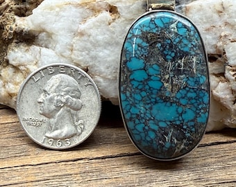 May include: Oval turquoise pendant with a silver-coloured metal frame and bail. The turquoise stone features a mottled pattern of blue and black. A quarter coin from 1963 is next to the pendant.