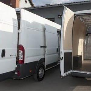 May include: A white cargo van with the rear doors open, revealing an empty cargo space. The van has a wooden interior and a black trim around the doors.
