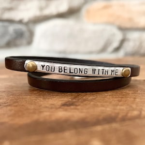 May include: A brown leather wrap bracelet with a silver metal plate that reads "YOU BELONG WITH ME". The bracelet has two brass rivets.