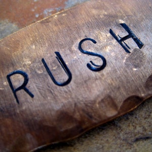 May include: Close-up of a copper metal tag with the word "RUSH" engraved in black, bold letters. The tag has a hammered texture and a warm, reddish-brown color. The background is a neutral, textured surface.