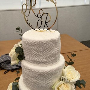 Könnte beinhalten: Eine dreistöckige weiße Hochzeitstorte mit einem spitzem Muster. Die oberste Etage ist mit einem goldenen Topper verziert, auf dem die Buchstaben "R & R" und das Datum "22.06.2023" stehen. Die Torte ist mit weißen Rosen und Eukalyptus verziert.