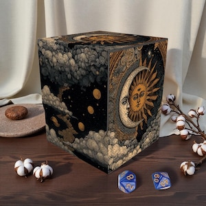 May include: A cube-shaped box with a celestial design featuring a sun and moon. The box is decorated with clouds, stars, and gold accents. Two blue dice are on the wooden surface in front of the box.