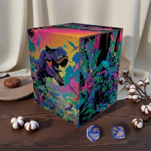 Puede incluir: Una caja de almacenamiento cúbica con un diseño colorido de dinosaurios y selva. La caja presenta una ilustración de T-Rex en tonos morados, azules y amarillos. El fondo incluye un follaje verde y negro vibrante. Dos dados azules están frente a la caja.