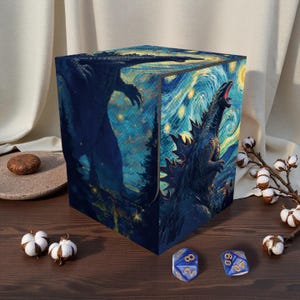 May include: A cube-shaped storage box featuring a stylized illustration of Godzilla against a starry night sky. The artwork is rendered in shades of blue and yellow, with a textured, painted appearance. Two blue dice are visible in the foreground.