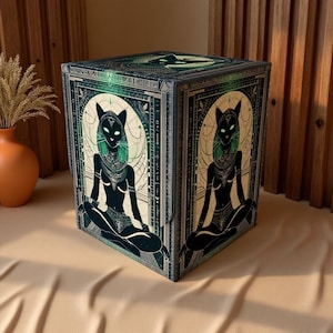 May include: A decorative cube with an Egyptian theme. The cube's sides showcase a seated figure with cat ears, green eyes, and detailed jewellery. The colour scheme includes black, green, and beige, with a geometric border.