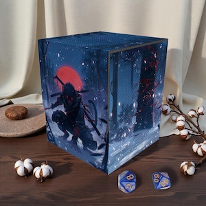 Op de afbeelding: Een kubusvormige dobbelsteenbox met een digitale kunstprint van een ninja in een besneeuwde bosscène met een rode zon. De doos is donkerblauw met een textuur. Twee blauwe en gouden dobbelstenen zijn zichtbaar.