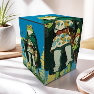 Könnte beinhalten: Würfelförmige dekorative Box mit einer Cartoon-Illustration eines Frosches in Ritterrüstung. Der Frosch trägt einen grünen Umhang und hält ein Schwert vor einem blauen Himmel mit gelben und weißen Wolken.