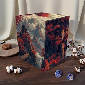 May include: A cube-shaped storage box featuring a scenic landscape with a pagoda, bridge, and red-leafed trees. The box has a mountain range backdrop with a cloudy sky. Two blue dice with white numbers are on a wooden surface.