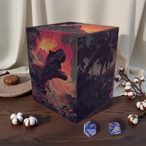 Puede incluir: Una caja de almacenamiento cúbica con una ilustración detallada de un Tyrannosaurus Rex en un entorno de jungla prehistórica. La obra de arte incluye una puesta de sol ardiente y un follaje exuberante. Se ven dos dados azules.