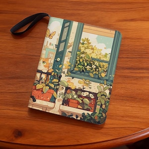 Può includere: Custodia rettangolare illustrata con un gatto che guarda da una finestra con piante e una farfalla. Ha un cinturino nero e poggia su una superficie di legno. L'illustrazione utilizza tonalità di verde, arancione e blu.