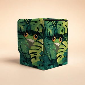 May include: A square storage box featuring a vibrant jungle pattern with large green monstera leaves and two illustrated frogs with prominent orange eyes peeking out from the foliage. The box has a structured design with a hinged lid.