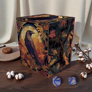 Puede incluir: Una caja de dados cúbica con una ilustración de un cuervo. La caja presenta un diseño de estilo vidriera con hojas de otoño en tonos naranja, amarillo y marrón. Se ven dos dados azules con números blancos.