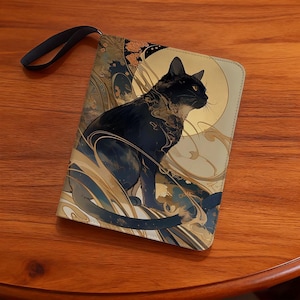 Puede incluir: Bolso de mano rectangular con una ilustración de un gato negro. El diseño incluye remolinos dorados y azul marino y una luna dorada. El bolso tiene una correa de muñeca negra. El gato es el foco central, sobre un fondo crema.