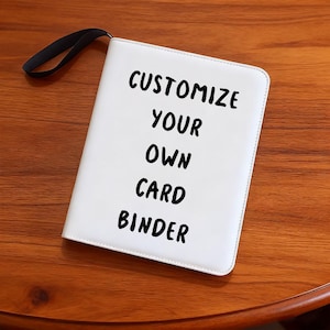 Puede incluir: Un archivador de tarjetas blanco con un asa negra y el texto "CUSTOMIZE YOUR OWN CARD BINDER" en negro. El archivador está sobre una superficie de madera.