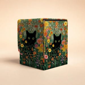 May include: A square storage box featuring a vibrant floral print with numerous colorful flowers in shades of orange, yellow, pink, and purple. Two black cats with bright yellow eyes are positioned among the dense green foliage and blossoms.