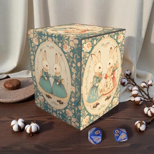 Puede incluir: Una caja cúbica con un estampado floral turquesa e ilustraciones ovaladas de conejos con vestidos. La caja tiene una estética vintage, con dos dados azules y ramitas de algodón cerca. La caja es probablemente para almacenamiento o exhibición.