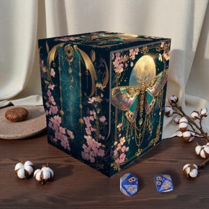 May include: A dark teal box with a butterfly and floral design. The butterfly has iridescent wings and is centered on the front of the box. Pink flowers and gold accents decorate the sides. Two blue dice are in front of the box.