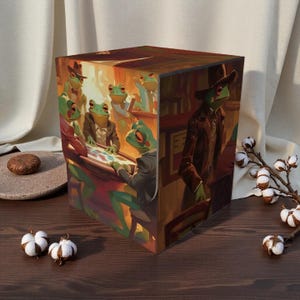 May include: A decorative cube featuring a whimsical illustration of anthropomorphic frogs in a casino setting. The artwork depicts frogs in suits and hats, playing cards and socializing in a dimly lit room. The cube is set on a dark wooden surface.