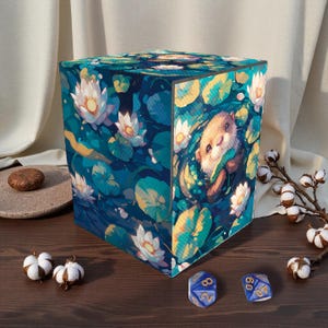 May include: A cube-shaped storage box featuring an illustration of an otter in a pond with lily pads and flowers. The box is primarily blue and green, with white and yellow accents. Two blue dice with gold numbers are visible.