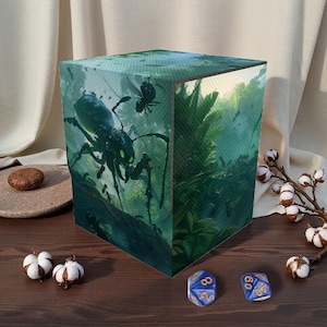 Könnte beinhalten: Eine würfelförmige Aufbewahrungsbox mit einer Fantasy-Kunstszene eines riesigen Insekts mit leuchtend grünen Augen in einer Dschungelumgebung. Die Box ist von Baumwollblüten und zwei blauen Würfeln umgeben.