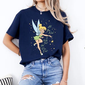 May include: Navy blue t-shirt with a cartoon image of Tinkerbell and the words "All You Need Is Faith Trust And Pixie Dust". The design includes stars and a fairy. A casual wear item.