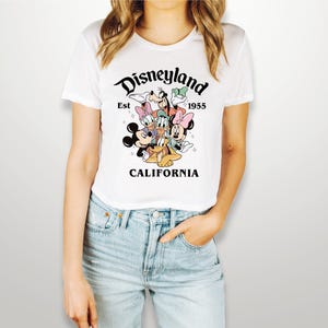 May include: White t-shirt with a graphic of Mickey Mouse, Minnie Mouse, Donald Duck, Goofy, and Pluto. The graphic is surrounded by the text "Disneyland Est 1955 California".