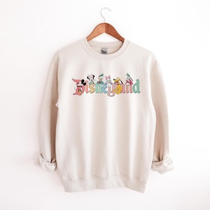 May include: A cream colored sweatshirt with a colorful graphic of Disney characters and the text "Disneyland" in a vintage font.