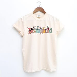 May include: A cream colored t-shirt with a vintage-style Disneyland logo featuring Mickey Mouse, Minnie Mouse, Donald Duck, Goofy, Pluto, and a few other characters. The logo is in rainbow colors.