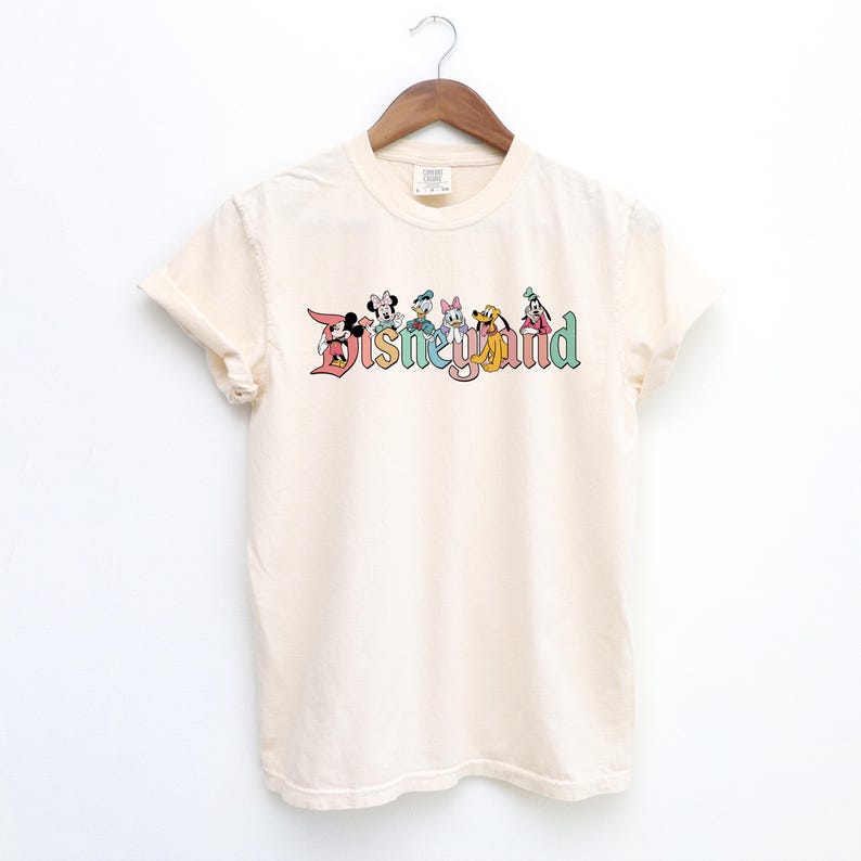 May include: A cream colored t-shirt with a vintage-style Disneyland logo featuring Mickey Mouse, Minnie Mouse, Donald Duck, Goofy, Pluto, and a few other characters. The logo is in rainbow colors.