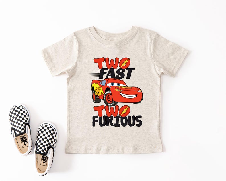 Peut inclure: Un t-shirt gris clair avec une voiture de course rouge de dessin anim&eacute; et le texte "TWO FAST TWO FURIOUS" en noir et rouge. Une paire de chaussures &agrave; enfiler &agrave; carreaux noirs et blancs est &agrave; gauche.
