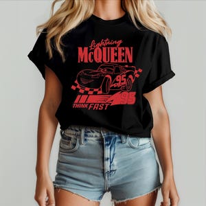 Może przedstawiać: Czarny T-shirt z czerwoną grafiką Błyskawicy McQueena z filmu Auta. Grafika zawiera tekst "Lightning McQueen", "Think Fast" i numer "95".