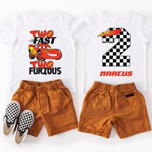 Peut inclure: Deux t-shirts blancs avec des motifs de voiture de course et le texte "Two Fast Two Furious" et un num&eacute;ro 2 &agrave; damier avec le nom "Marcus". Ils sont assortis &agrave; des shorts marron et des chaussures &agrave; enfiler &agrave; carreaux.