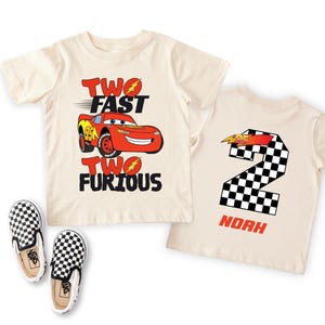Peut inclure: Deux t-shirts cr&egrave;me sur le th&egrave;me de Cars. L'un pr&eacute;sente Flash McQueen et le texte "TWO FAST TWO FURIOUS". L'autre affiche un num&eacute;ro 2 &agrave; damier et le nom "NOAH". Des chaussures &agrave; enfiler &agrave; carreaux noirs et blancs sont &eacute;galement visibles.