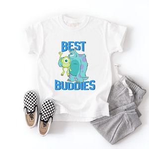 Peut inclure: T-shirt blanc avec l'inscription bleue "BEST BUDDIES" et un dessin animé de deux monstres. L'image comprend un pantalon de survêtement gris et des chaussures à enfiler à carreaux noirs et blancs.