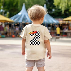 Peut inclure: Un t-shirt beige pour enfant avec un grand chiffre 2 au dos, orn&eacute; d'un motif &agrave; carreaux noir et blanc et d'un motif de flamme rouge et jaune. L'enfant porte un short gris.
