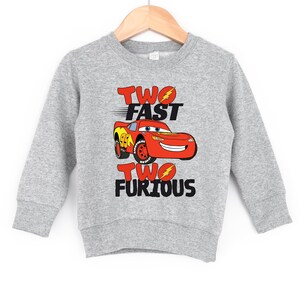 Peut inclure: Un sweat-shirt gris avec l'inscription "TWO FAST TWO FURIOUS" en rouge et noir. Une voiture de course rouge de dessin anim&eacute; avec le num&eacute;ro 94 est au centre. Le sweat-shirt est suspendu &agrave; un cintre en bois.