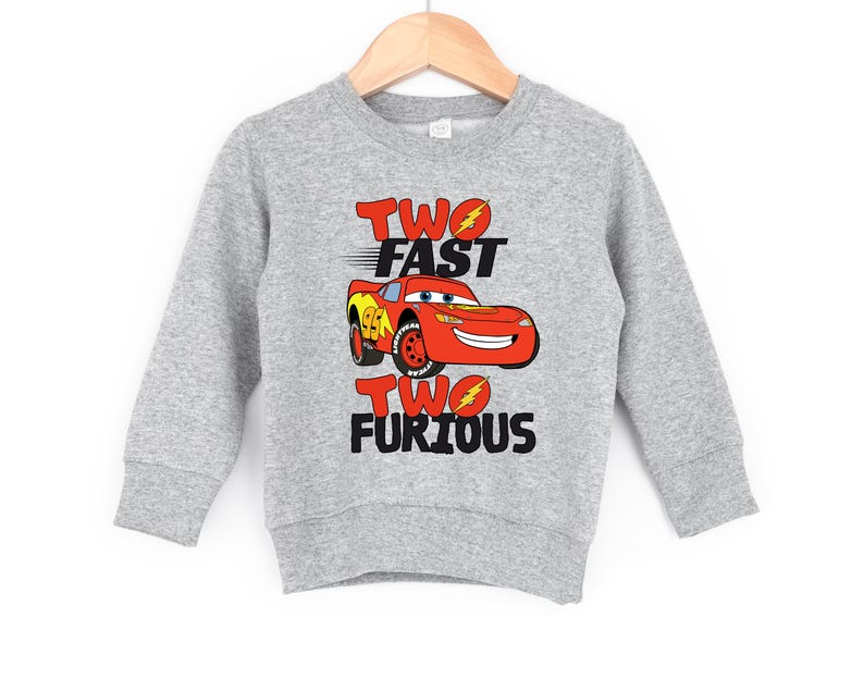 Peut inclure: Un sweat-shirt gris avec l'inscription "TWO FAST TWO FURIOUS" en rouge et noir. Une voiture de course rouge de dessin anim&eacute; avec le num&eacute;ro 94 est au centre. Le sweat-shirt est suspendu &agrave; un cintre en bois.