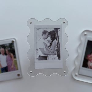 May include: Clear acrylic photo frames with a wavy edge design. Each frame holds a small black and white photograph. The frames are displayed on a white surface. The photos appear to be of a couple.