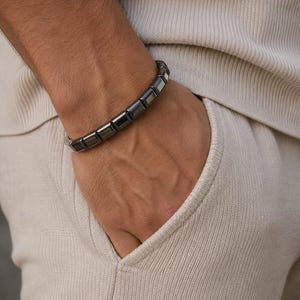May include: A dark grey metallic bracelet composed of a series of rectangular, polished links. The bracelet features a smooth, reflective surface and a segmented design that wraps around the wrist, showcasing a uniform, sleek, and industrial appearance.